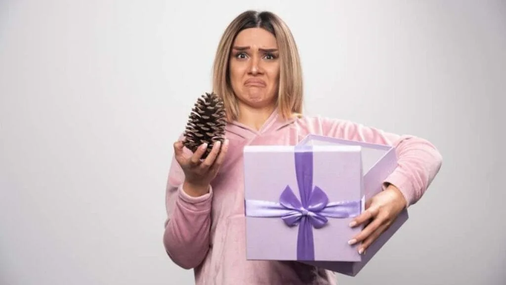 Mulher com expressão de decepção segurando uma pinha e uma caixa de presente